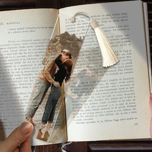 May include: A clear acrylic bookmark featuring a printed photograph of a couple embracing and kissing on a beach. The rectangular bookmark has a rounded top with a hole, through which a cream-colored decorative tassel is attached with a knotted cord.