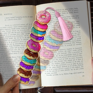 Op de afbeelding: Een kleurrijke boekenlegger met een stapel geïllustreerde donuts met roze glazuur en hagelslag. De boekenlegger heeft een roze kwast en staat in een open boek.