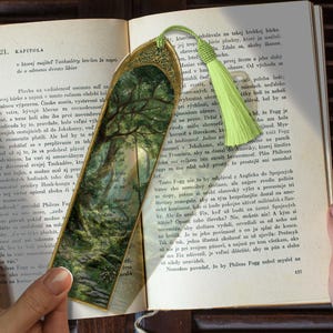 Può includere: Segnalibro decorativo con un'illustrazione di una scena di foresta. Il segnalibro ha una parte superiore in filigrana dorata con un nappa verde.
