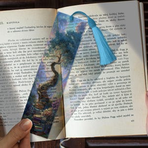 May include: A decorative bookmark with a blue tassel, resting on an open book. The bookmark features a watercolor painting of a tree with a winding trunk and a stack of books.