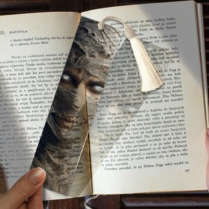 May include: A bookmark with a mummy design and a cream tassel. The mummy's face, with white eyes and bandages, is visible. The bookmark is on a book with text, likely a novel.