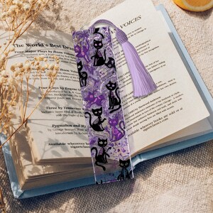 May include: A clear acrylic bookmark featuring a repeating pattern of black cat skeletons and purple cat silhouettes. The bookmark has a lavender tassel and is placed on an open book. The book's pages are visible.