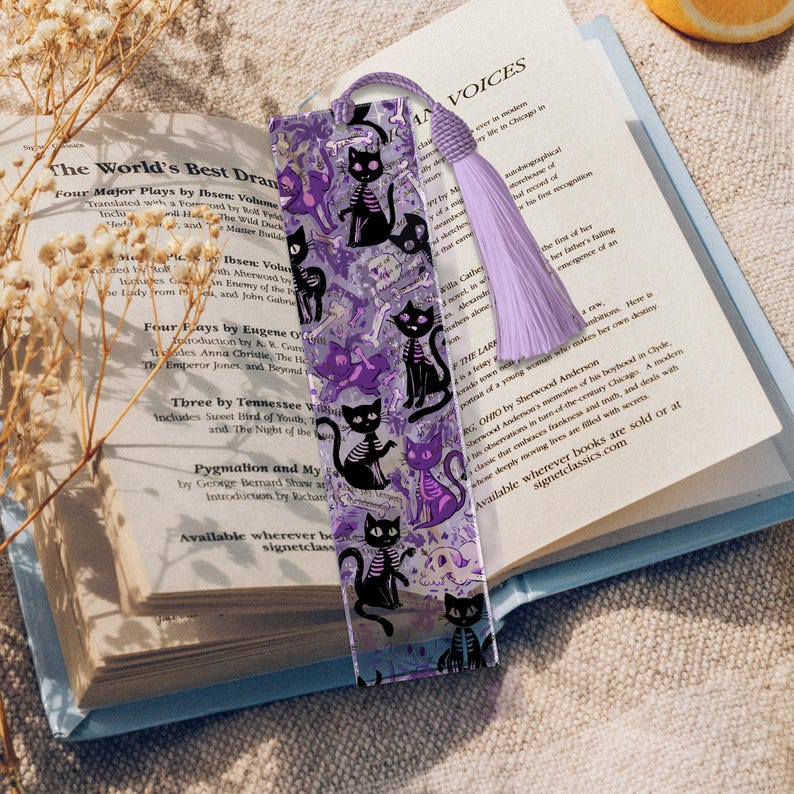 May include: A clear acrylic bookmark featuring a repeating pattern of black cat skeletons and purple cat silhouettes. The bookmark has a lavender tassel and is placed on an open book. The book's pages are visible.