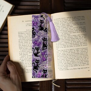 May include: A bookmark featuring a repeating pattern of black skeleton cats and purple floral elements. The bookmark has a lavender tassel and is placed in an open book.