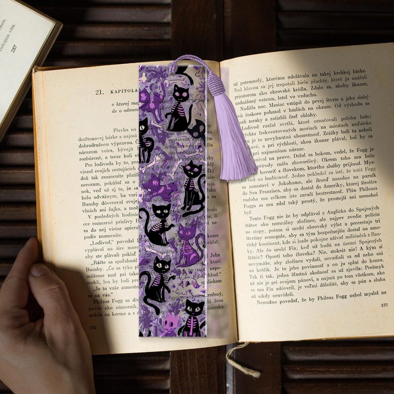May include: A bookmark featuring a repeating pattern of black skeleton cats and purple floral elements. The bookmark has a lavender tassel and is placed in an open book.