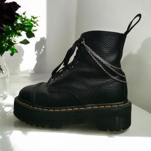 Könnte beinhalten: Schwarze Leder-Plateau-Stiefel mit silbernem Kettendetail. Die Stiefel haben eine dicke Plateau-Sohle und einen Schnürverschluss.