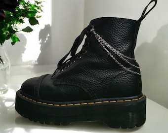 Puede incluir: Botas de plataforma de cuero negro con un detalle de cadena plateada. Las botas tienen una suela de plataforma gruesa y un cierre de cordones.