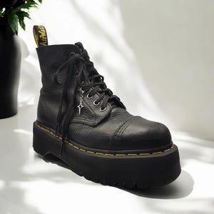 Könnte beinhalten: Schwarze Plateau-Stiefel aus Leder mit gelben Nähten und einem silbernen Anhänger. Die Stiefel haben eine dicke Plateau-Sohle und einen Schnürverschluss.