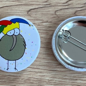 Könnte beinhalten: Ein runder Button mit einem Cartoon-Kiwi-Vogel, der einen Narrenhut in Rot, Gelb, Grün und Blau trägt. Der Button hat einen weißen Hintergrund mit bunten Konfetti. Die Rückseite des Buttons ist silberfarben mit einer Nadel.