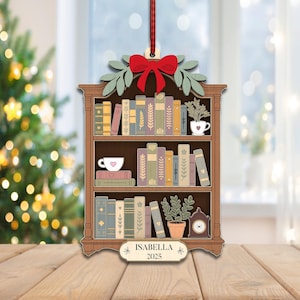 May include: Wooden Christmas tree ornament shaped like a bookshelf, with a red bow, green leaves, and a red ribbon. The bookshelf is filled with books, a teacup, a plant, and a clock. The name "ISABELLA" and the year "2025" are displayed.