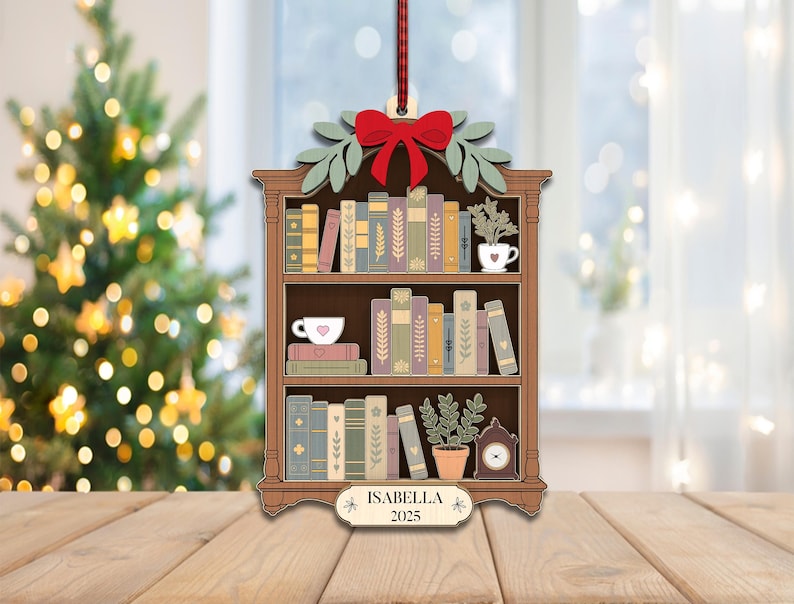 May include: Wooden Christmas tree ornament shaped like a bookshelf, with a red bow, green leaves, and a red ribbon. The bookshelf is filled with books, a teacup, a plant, and a clock. The name "ISABELLA" and the year "2025" are displayed.