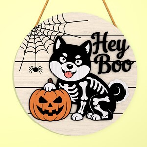 Może przedstawiać: Okrągły drewniany znak na Halloween z czarno-białym psem Shiba Inu w kostiumie szkieletu trzymającym pomarańczową dynię. Znak zawiera napis "Hey Boo" i wz&oacute;r pajęczyny.