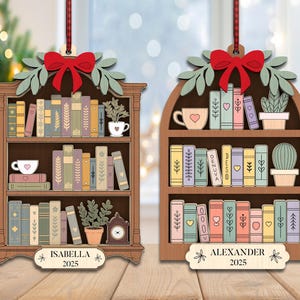 May include: Wooden bookshelf Christmas decorations with red bows, green leaves, and colourful books. One ornament says "Isabella 2025" and the other says "Alexander 2025". Each shelf has a teacup and a plant.