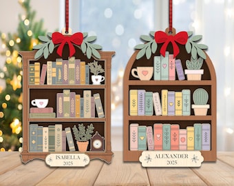 May include: Wooden bookshelf Christmas ornaments with red bows, green leaves, and colorful books. One ornament says "Isabella 2025" and the other says "Alexander 2025". Each shelf has a teacup and a plant.