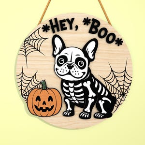 Może przedstawiać: Okrągły drewniany znak na Halloween z motywem szkieletu buldoga francuskiego, dyni i pajęczyn. Znak głosi "Hey, Boo" czarnymi literami i wisi na naturalnej lince.