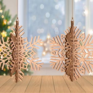 May include: Two wooden snowflake ornaments hanging from strings. The snowflakes are intricately cut with a natural wood finish. A Christmas tree with blurred lights is in the background, suggesting a holiday theme. The ornaments are displayed on a wooden surface.