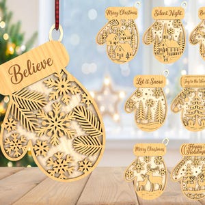 May include: Wooden Christmas ornaments in the shape of mittens. One ornament says "Believe" and features snowflake and leaf cutouts. Other ornaments have holiday phrases like "Merry Christmas" and "Silent Night" with winter scene cutouts.