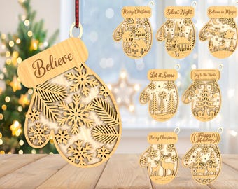Op de afbeelding: Een collectie lasergesneden houten wantvormige ornamenten met winterse taferelen en tekst. De ontwerpen bevatten sneeuwvlokken, dennentakken, rendieren, een kerk en een arrenslee. Teksten zijn onder andere Believe, Merry Christmas, Silent Night, Believe in Magic, Let it Snow, Joy to the World en Happy Holidays.