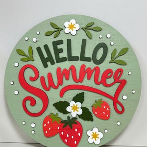 K&ouml;nnte beinhalten: Runder Holzschild mit hellgr&uuml;nem Hintergrund, auf dem die Worte "HELLO Summer" in Gr&uuml;n und Rot stehen. Das Schild ist mit roten Erdbeeren, wei&szlig;en Blumen und gr&uuml;nen Bl&auml;ttern verziert.