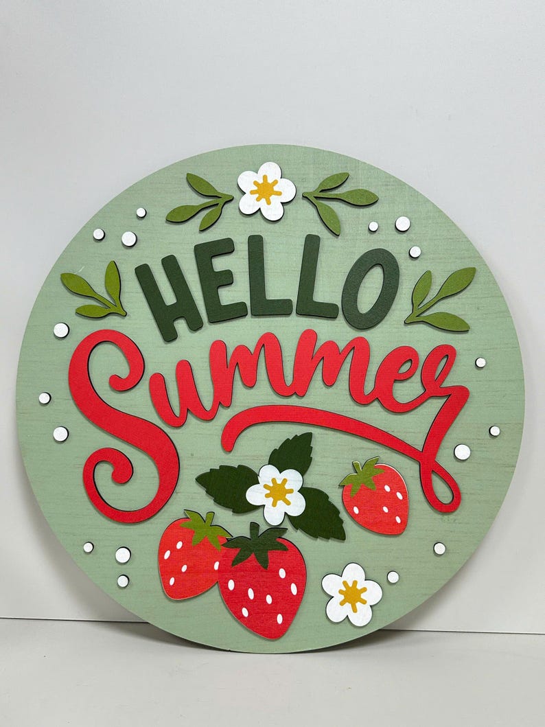 K&ouml;nnte beinhalten: Runder Holzschild mit hellgr&uuml;nem Hintergrund, auf dem die Worte "HELLO Summer" in Gr&uuml;n und Rot stehen. Das Schild ist mit roten Erdbeeren, wei&szlig;en Blumen und gr&uuml;nen Bl&auml;ttern verziert.