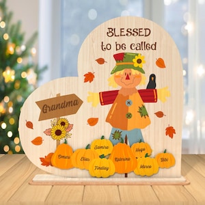 May include: A wooden heart-shaped sign with the text "Blessed to be called Grandma" and a scarecrow illustration. The sign has a brown arrow pointing to the text "Grandma" and a row of pumpkins with names written on them.