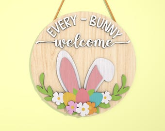 Pode incluir: Placa de Páscoa redonda de madeira com o texto "EVERY - BUNNY welcome". A placa apresenta orelhas de coelho, ovos de Páscoa coloridos e detalhes florais. É projetada com um fundo de grão de madeira clara e uma corda de juta para pendurar.