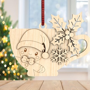 May include: Wooden Christmas ornament shaped like a cup with a gingerbread person wearing a Santa hat. The cup design includes holly leaves and a snowflake. A red and black plaid ribbon is attached for hanging. The ornament is made of light-coloured wood.