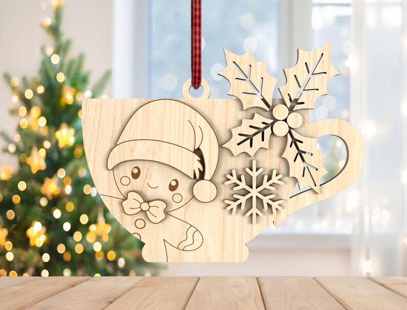 May include: Wooden Christmas ornament shaped like a cup with a gingerbread person wearing a Santa hat. The cup design includes holly leaves and a snowflake. A red and black plaid ribbon is attached for hanging. The ornament is made of light-coloured wood.