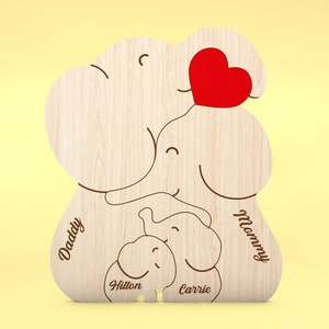 Op de afbeelding: Houten familie-olifantvormige decoratie met gegraveerde namen en een rood hart. Het ontwerp bevat de woorden "Daddy", "Mommy", "Hilton" en "Carrie". De olifantenfamilie is gemaakt van lichtgekleurd hout.