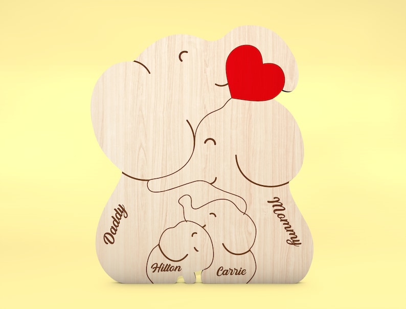 Op de afbeelding: Houten familie-olifantvormige decoratie met gegraveerde namen en een rood hart. Het ontwerp bevat de woorden "Daddy", "Mommy", "Hilton" en "Carrie". De olifantenfamilie is gemaakt van lichtgekleurd hout.