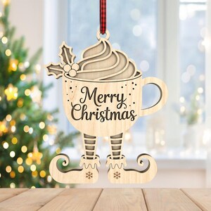 May include: Wooden Christmas ornament shaped as a coffee cup with elf legs. The cup has whipped cream and holly on top, and the words "Merry Christmas" are written on the front. The ornament hangs from a red and black checkered ribbon.