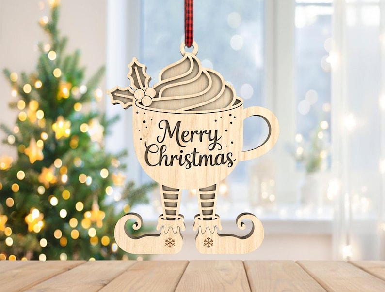May include: Wooden Christmas ornament shaped as a coffee cup with elf legs. The cup has whipped cream and holly on top, and the words "Merry Christmas" are written on the front. The ornament hangs from a red and black checkered ribbon.