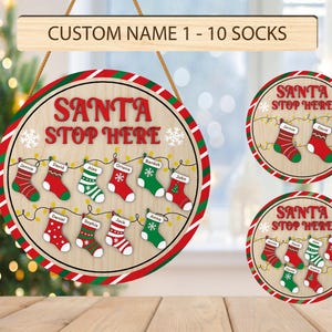 May include: A set of three round wooden signs with a red and white striped border. Each sign has the text "Santa Stop Here" in red letters and features a white snowflake and a string of lights with red and green Christmas stockings. The stockings have names written on them.