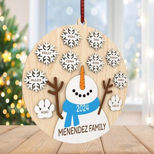 May include: A round wooden ornament with a snowman in the center. The snowman is wearing a blue scarf and a hat with the year 2024 on it. The ornament is decorated with snowflakes and paw prints with names on them. The text "MENENDEZ FAMILY" is at the bottom of the ornament.