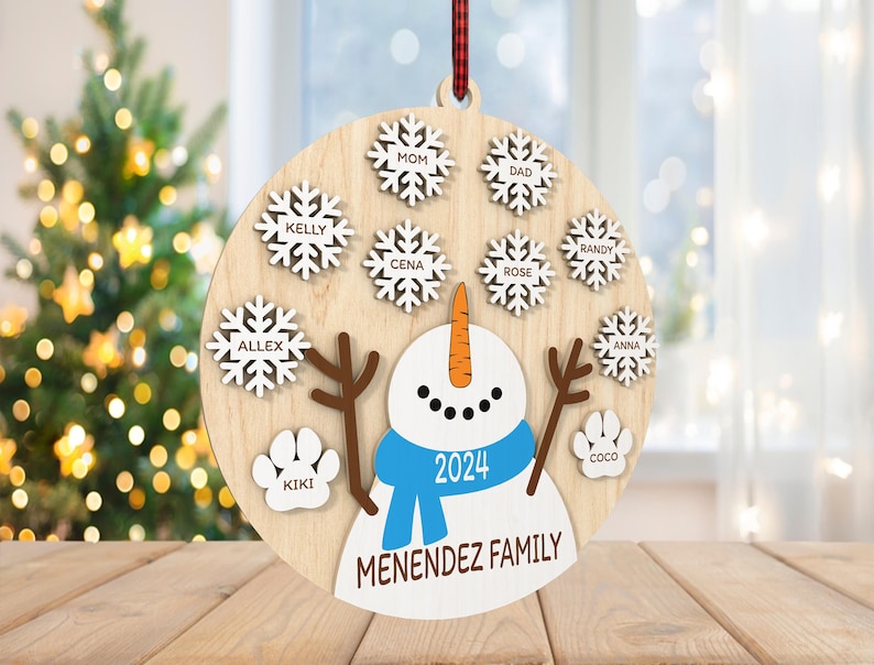 May include: A round wooden ornament with a snowman in the center. The snowman is wearing a blue scarf and a hat with the year 2024 on it. The ornament is decorated with snowflakes and paw prints with names on them. The text "MENENDEZ FAMILY" is at the bottom of the ornament.