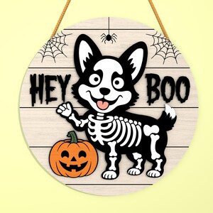 Może przedstawiać: Okrągły drewniany znak na Halloween z kresk&oacute;wkowym szkieletem corgi i dynią. Znak głosi "HEY BOO" czarnymi literami, z pajęczynami i pająkiem na g&oacute;rze. Znak jest zawieszony na brązowej linie.