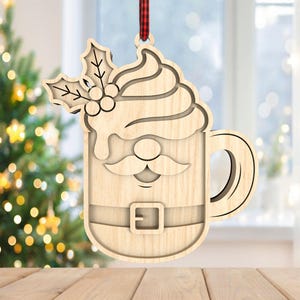 May include: Wooden Christmas ornament in the shape of a Santa-themed coffee mug. The ornament features a Santa face, belt, and whipped cream design, with holly leaves and berries at the top. It hangs from a red and black plaid ribbon.