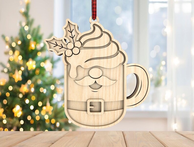 May include: Wooden Christmas ornament in the shape of a Santa-themed coffee mug. The ornament features a Santa face, belt, and whipped cream design, with holly leaves and berries at the top. It hangs from a red and black plaid ribbon.