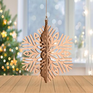 May include: A wooden snowflake ornament with intricate cut-out details hangs from a string. The natural wood color contrasts with the blurred background of a Christmas tree and a window with soft, white curtains. The ornament is a festive holiday decoration.