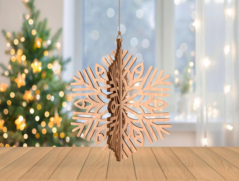 May include: A wooden snowflake ornament with intricate cut-out details hangs from a string. The natural wood color contrasts with the blurred background of a Christmas tree and a window with soft, white curtains. The ornament is a festive holiday decoration.
