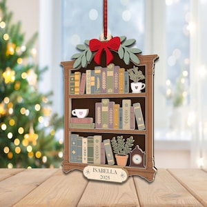 May include: Wooden Christmas tree ornament shaped like a bookshelf, with a red bow, green leaves, and a hanging ribbon. The ornament depicts a bookshelf with books, plants, and a clock. The name "Isabella" and the year "2025" are displayed on a small sign.