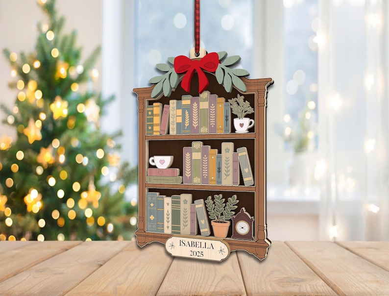 May include: Wooden Christmas tree ornament shaped like a bookshelf, with a red bow, green leaves, and a hanging ribbon. The ornament depicts a bookshelf with books, plants, and a clock. The name "Isabella" and the year "2025" are displayed on a small sign.