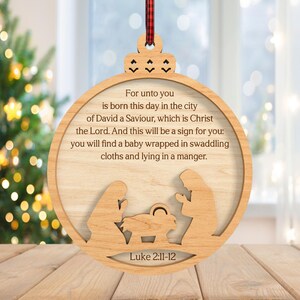 May include: Wooden Christmas ornament with a circular design. The ornament features a cutout nativity scene and the text "For unto you is born this day..." from Luke 2:11-12. A red and black plaid ribbon is attached for hanging.