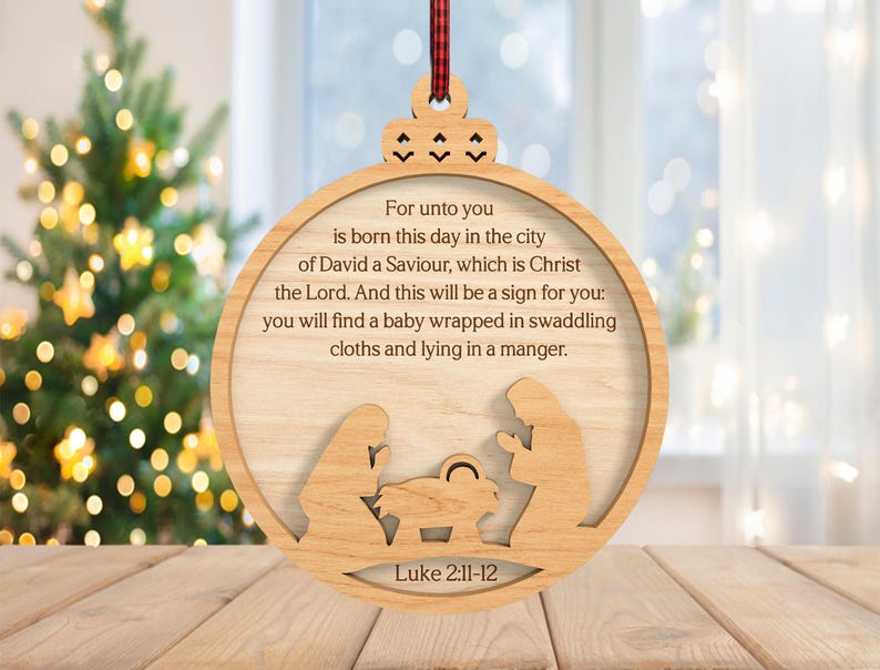 May include: Wooden Christmas ornament with a circular design. The ornament features a cutout nativity scene and the text "For unto you is born this day..." from Luke 2:11-12. A red and black plaid ribbon is attached for hanging.