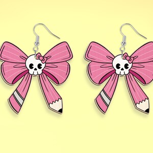 May include: A pair of pink bow earrings with a skull charm and a small pink bow on the skull. The bow has a pencil-shaped detail on the bottom.
