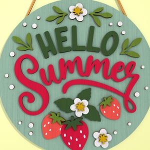 K&ouml;nnte beinhalten: Runder Holzschild mit den Worten "HELLO Summer" in gr&uuml;ner und roter Schrift. Das Schild zeigt Erdbeeren, wei&szlig;e Blumen und gr&uuml;ne Bl&auml;tter. Ein beiges Seil ist zum Aufh&auml;ngen befestigt. Der Hintergrund ist hellt&uuml;rkis.