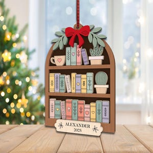 May include: Wooden bookshelf Christmas ornament with a red bow, green leaves, and a red ribbon. The bookshelf is filled with colourful books, a mug, and a cactus. The name "ALEXANDER" and the year "2025" are engraved on a small wooden sign.