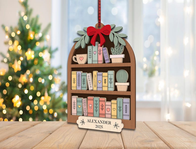 May include: Wooden bookshelf Christmas ornament with a red bow, green leaves, and a red ribbon. The bookshelf is filled with colourful books, a mug, and a cactus. The name "ALEXANDER" and the year "2025" are engraved on a small wooden sign.