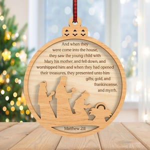 May include: Wooden Christmas ornament with a circular design. The ornament features a cutout nativity scene and text from Matthew 2:11. The text reads "And when they were come into the house...gifts; gold, and frankincense, and myrrh."
