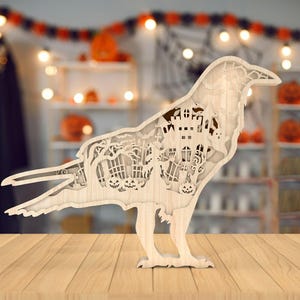 Peut inclure: Une découpe en bois d'un corbeau avec un motif de maison hantée à l'intérieur. Le corbeau est posé sur une surface en bois.